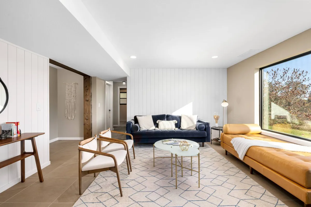 A contemporary and cozy lounge area featuring a blue velvet sofa, tan chaise lounge, decorative coffee table, and a geometric patterned rug. Natural light streams in through a large window, inviting guests to relax, read, or socialize while enjoying views of the outdoors.