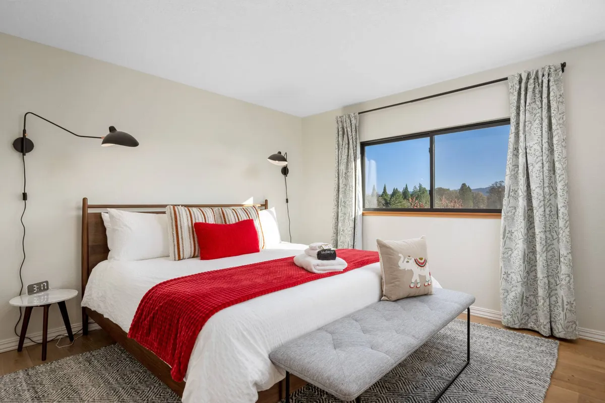 A tastefully decorated king bedroom with a bed featuring a red throw blanket and decorative pillows, a bench at the foot of the bed, wall-mounted lights, and a window with a view. The room is bathed in natural light, enhancing its cozy and restful ambiance.