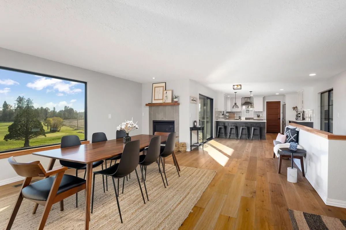 A warm, open interior featuring a spacious dining table set near a large window, with the adjacent kitchen and a cozy fireplace visible. The window frames a beautiful sunset, inviting the outdoors inside and setting the scene for gatherings and special meals.