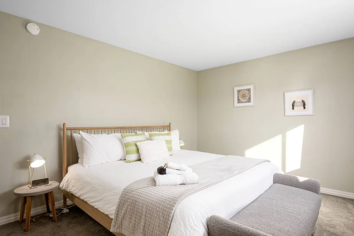 A serene bedroom featuring a comfortable bed adorned with decorative pillows, a nightstand with a lamp, and framed artwork on the wall. Natural light filters in, highlighting the cozy and welcoming atmosphere.