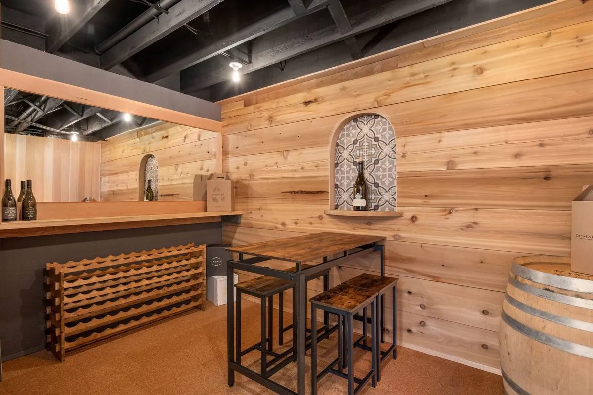 A warm, inviting bar area with a wooden bar, metal stools, and wine barrels set against a decorative arched wall. The space is ideal for wine tasting or casual gatherings, capturing the ambiance of private wine enjoyment.