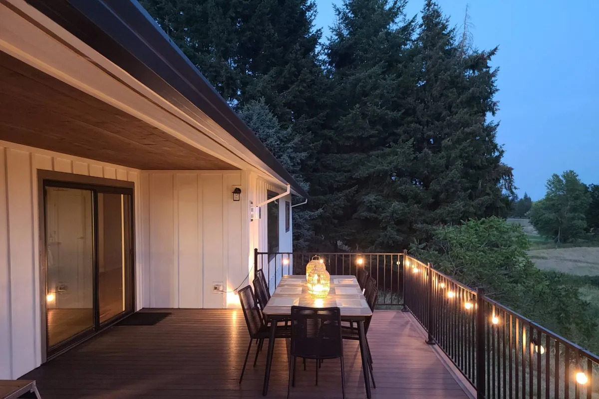 An outdoor dining area on a deck, complete with a dining table and chairs, decorative lantern, and surrounded by lush trees and a sturdy railing. The setting is serene and cozy, ideal for enjoying meals or socializing in the fresh air.